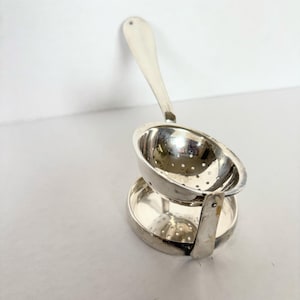 May include: A silver tea infuser with a handle and a perforated bowl, resting on a matching saucer-like base. The infuser is designed to hold loose tea leaves for steeping. The design is simple and elegant.