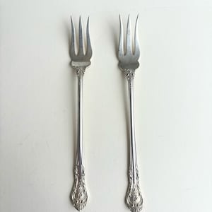 May include: Two silver-toned metal forks with three tines each. The handles feature an ornate, embossed floral pattern at the base and a smooth, slender neck leading to the tines. The metal has a polished, reflective finish throughout.