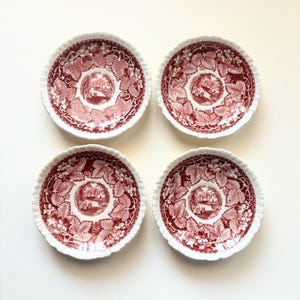 May include: A set of four round ceramic saucers featuring a deep red transferware pattern. Each saucer displays a central landscape scene surrounded by a detailed border of grape leaves and vines on a white background with scalloped edges.
