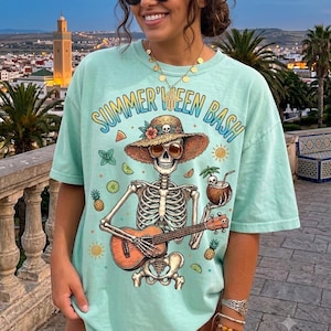 May include: A light mint green t-shirt featuring a graphic print of a skeleton wearing a straw hat and sunglasses, playing a ukulele while holding a coconut drink. The text "SUMMER'WEEN BASH" is printed in blue and yellow above the skeleton.