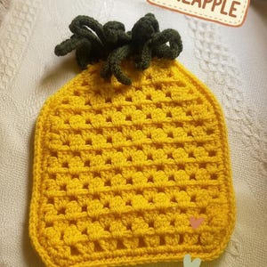 May include: A yellow crocheted square with a textured grid pattern and a dark green crocheted top resembling pineapple leaves. A rectangular label with a dashed border contains the text PINEAPPLE in brown capital letters at the top right.