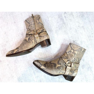 May include: A pair of ankle boots featuring a snakeskin pattern in shades of beige, gray, and black. The boots have a low block heel, a side zipper closure, and a decorative strap across the ankle secured with a metallic ring and studs.