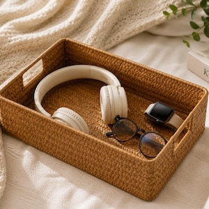 May include: A rectangular woven rattan storage tray with integrated side handles. Inside the tray are a pair of off-white over-ear headphones, a pair of black-rimmed eyeglasses, and a digital smartwatch with a beige band and a black screen.