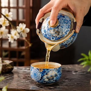 May include: A ceramic teapot and matching teacup featuring a blue and white floral pattern with gold accents. The teapot has a scalloped lid and a dark brown woven handle, while the teacup is filled with amber-coloured tea being poured.