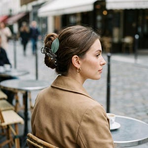May include: A hair claw clip featuring a green leaf-shaped accent and a silver-toned butterfly design with small white pearl-like beads. The clip is secured in a brown hair bun, complemented by a tan coat and small gold hoop earrings.
