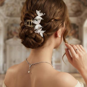 May include: A clear plastic hair claw clip adorned with white fabric butterfly embellishments and small pearl accents, secured in a braided updo hairstyle, paired with a silver-toned necklace featuring a central teardrop pendant and a ring.