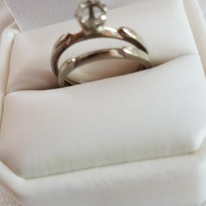 May include: A silver engagement ring with a clear, round-cut gemstone set in a raised prong setting. The ring is displayed in a white ring box, showcasing its elegant design. The band has a split shank design.