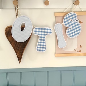 May include: A fabric banner spelling the name Otis, featuring padded letters in alternating light blue pinstripe and blue-and-white gingham check patterns, each outlined with white scalloped trim and suspended by white string.