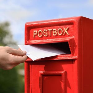 May include: A bright red metal postbox with the word POSTBOX embossed in gold lettering across the top. A white rectangular envelope is being inserted into the horizontal mail slot located directly beneath the embossed text on the front.