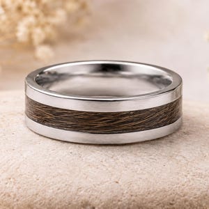 Op de afbeelding: Een zilverkleurige metalen ring met een centrale inleg van donkerbruin, getextureerd organisch materiaal. De band heeft een gladde, gepolijste afwerking aan de randen, die het vezelige, haarachtige uiterlijk van de inleg rondom omlijst.