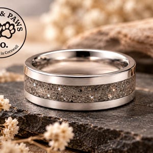 Op de afbeelding: Een zilverkleurige metalen ring met een centrale inleg van gestructureerd grijs zand met kleine sprankelende spikkels. De band heeft gepolijste randen en een gladde afwerking. Een rond logo linksboven vermeldt Shell & Paws Co. Handmade in Cornwall.