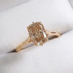 May include: A gold ring featuring a rectangular, champagne-coloured gemstone set in a four-prong setting. The band is smooth and polished, showcasing a warm metallic tone that complements the faceted, light brown centre stone held securely.