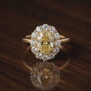 Op de afbeelding: Een geelgouden ring met een centrale ovaal geslepen gele edelsteen, omringd door een halo van twaalf kleine, ronde, helderwitte diamanten. De ringband is glad en gepolijst en toont een klassiek ontwerp met een levendige, warmgekleurde steen.