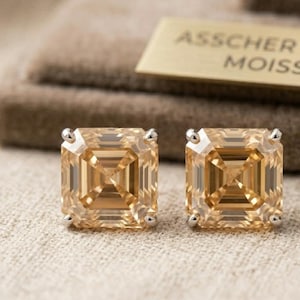 Può includere: Un paio di orecchini a lobo con moissanite color champagne a taglio Asscher quadrato, incastonati in griffe di metallo color argento. Sullo sfondo è visibile una piccola targhetta rettangolare dorata con la scritta ASSCHER CUT CH MOISSANITE.
