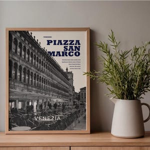 Könnte beinhalten: Ein gerahmter Wandkunstdruck mit einer Schwarz-Weiß-Fotografie der Piazza San Marco in Venedig, Italien. Der Druck enthält den fetten blauen Text "PIAZZA SAN MARCO" und "VENEZIA" in Weiß sowie weitere kleinere beschreibende Textdetails.
