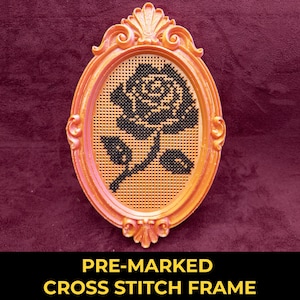 May include: An oval-shaped, orange-toned frame with ornate, carved detailing at the top and bottom. Inside the frame is a tan, perforated grid material featuring a black cross-stitch pattern of a single rose with a stem and two leaves.