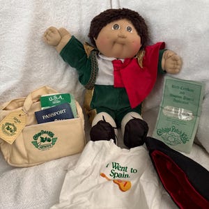 May include: A Cabbage Patch Kids doll with brown hair wearing a green and red outfit, accompanied by a beige Cabbage Patch Kids Airways travel bag, a passport, a white t-shirt with the text I Went to Spain, and a birth certificate document.