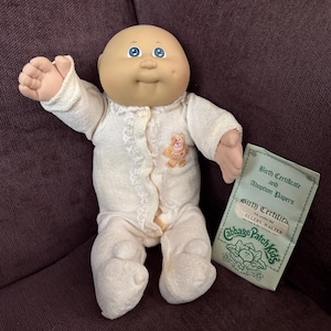 May include: A Cabbage Patch Kids doll with a bald head, blue eyes, and a dimpled face, wearing a cream-colored long-sleeved onesie with a small teddy bear patch and lace trim. Beside the doll is a paper document titled Birth Certificate and Adoption Papers.
