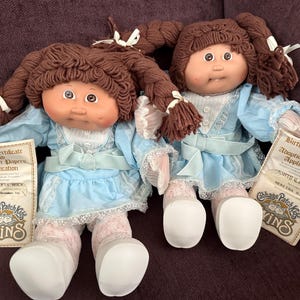 May include: Two Cabbage Patch Kids dolls with brown yarn hair in pigtails, wearing light blue dresses with white lace trim and white shoes. Each doll is accompanied by a paper birth certificate and adoption papers application labeled Twins.