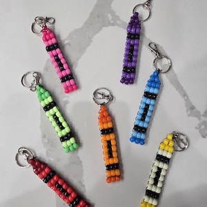 May include: Six crayon-shaped keychains crafted from colourful plastic pony beads. Each keychain features a distinct colour—pink, purple, light green, orange, blue, and yellow—accented with black beads and attached to a silver metal lobster clasp.