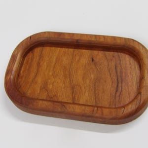 May include: A rectangular wooden tray with rounded corners and a recessed center. The tray features a warm, reddish-brown wood grain pattern with visible natural streaks and a smooth, polished finish throughout the entire surface area.