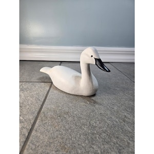 May include: A white carved wooden duck figurine with a smooth matte finish and a black beak. The bird is depicted in a resting position with its head turned slightly to the side, featuring a simple, clean design with no additional markings or details.