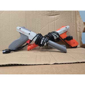 May include: Two Nintendo Zapper light gun controllers, one in grey and one in orange, featuring the Nintendo logo on the side. Both controllers are connected by a black coiled cord and include a black connector plug at the end of the cable.
