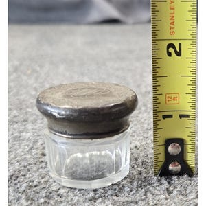 May include: A small, clear glass jar with a faceted base and a dark, tarnished metal lid featuring an engraved monogram. A yellow Stanley measuring tape is positioned next to the jar, indicating a height of approximately 1.25 inches for the item.