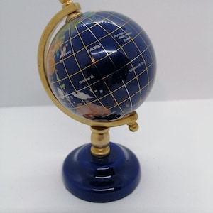 Könnte beinhalten: Ein dekorativer Globus mit einer tiefblauen Ozeanoberfläche und eingelegten Steinkontinenten. Der Globus ist auf einem goldfarbenen Metallmeridianbogen und einem runden blauen Sockel montiert. Weiße Beschriftungen zeigen Pacific, Tokyo, Honolulu und Pearl Harbor.