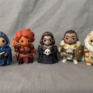 Könnte beinhalten: Ein Set aus fünf stilisierten Fantasy-Figuren. Die Gruppe besteht aus einer blau behuften Figur mit Buch, einer rothaarigen Figur mit Fackel, einer dunkel gekleideten Figur mit Totenkopf, einem Ritter in weiß-goldener Rüstung und einem Löwen.