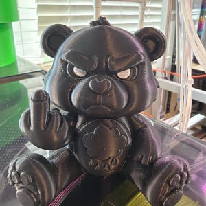 May include: A black 3D-printed bear figurine with a grumpy facial expression and white eyes. The bear is sitting down and holding up its middle finger on its right hand. A cloud-shaped emblem with raindrops is visible on the bear's chest.