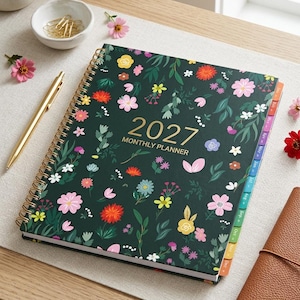 May include: A 2027 monthly planner with a dark green cover featuring a colorful floral pattern. The planner has gold spiral binding and gold text on the front. It includes side tabs for each month and is accompanied by a gold pen.