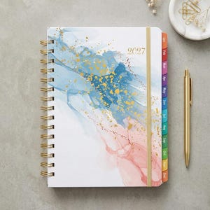 May include: A 2027 spiral-bound planner featuring a white cover with blue and pink watercolor-style abstract patterns and gold speckles. The planner includes a gold elastic closure, gold wire binding, and multicolored monthly tabs labeled Jan through Dec.