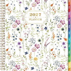 May include: A 2027 planner with a white cover featuring a colorful watercolor floral pattern of various wildflowers in shades of pink, purple, orange, blue, and yellow. The planner has a gold wire-o binding and monthly tabs along the right edge.