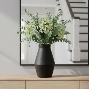 May include: A matte black ceramic vase with a tapered neck and a wide, rounded base. The vase is filled with a lush arrangement of light green hydrangea blossoms and slender eucalyptus stems with small, rounded leaves extending outward.