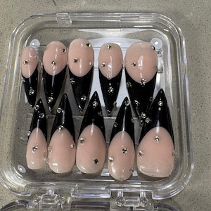 May include: A set of ten stiletto-shaped press-on nails arranged in a clear plastic storage case. Each nail features a glossy nude base with a sharp black French tip and is decorated with small, scattered silver metallic studs and clear rhinestones.