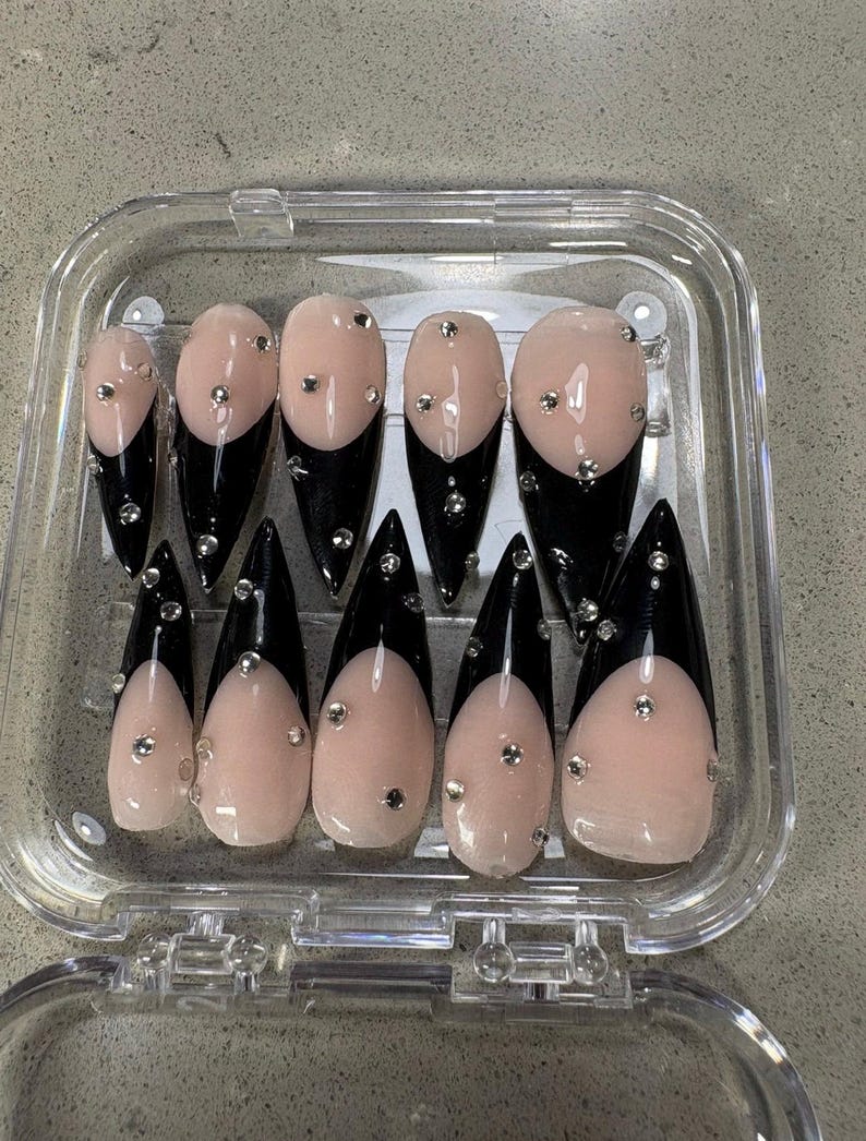 May include: A set of ten stiletto-shaped press-on nails arranged in a clear plastic storage case. Each nail features a glossy nude base with a sharp black French tip and is decorated with small, scattered silver metallic studs and clear rhinestones.