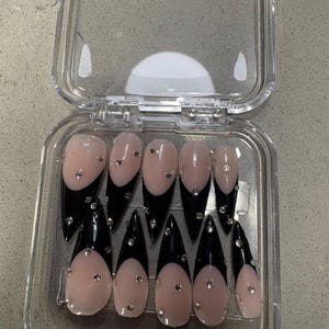 May include: A set of ten stiletto-shaped press-on nails arranged in a clear plastic storage case. Each nail features a nude base with a glossy black French tip and is embellished with small, clear, reflective rhinestones scattered across the surface.