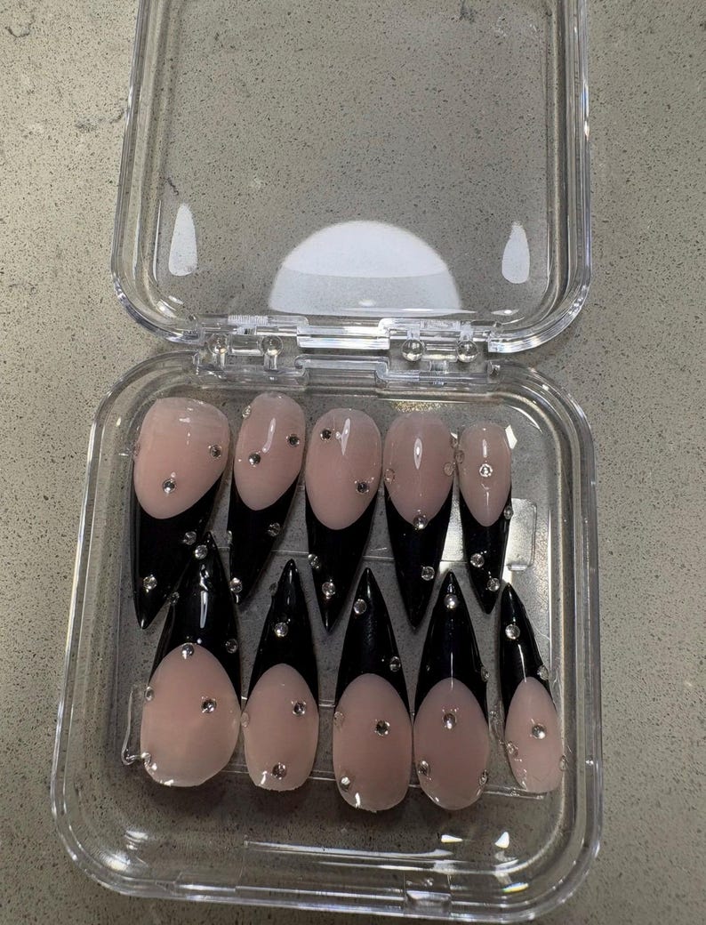 May include: A set of ten stiletto-shaped press-on nails arranged in a clear plastic storage case. Each nail features a nude base with a glossy black French tip and is embellished with small, clear, reflective rhinestones scattered across the surface.