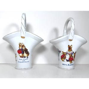 May include: Two white ceramic baskets with arched handles, each featuring colorful illustrations of Beatrix Potter characters. One basket depicts Peter Rabbit and Benjamin Bunny, while the other shows Mrs. Rabbit with small bunnies.