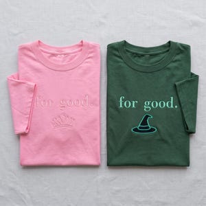 May include: A light pink t-shirt with the embroidered text for good. and a crown icon, alongside a dark green t-shirt with the embroidered text for good. and a witch hat icon. Both shirts feature short sleeves and a crew neckline design.