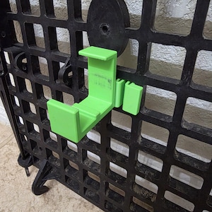 May include: A bright lime green plastic hook attachment mounted onto a black grid panel. The hook features a U-shaped cradle design with a rectangular base and a secondary smaller rectangular tab extending from the side, securely locked into the grid.