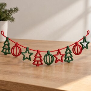 May include: A festive garland featuring a red cord strung with alternating green and red ornaments. The ornaments are shaped like stylized Christmas trees, round baubles, and stars, all with a simple, open-frame design and matte finish.