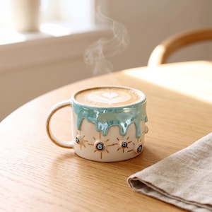 May include: A ceramic mug with a glossy light blue drip glaze on the upper rim and a white base. The exterior features a repeating pattern of small blue and white evil eye symbols surrounded by gold starburst lines and gold rim detailing.