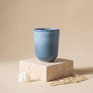 May include: A cylindrical ceramic cup featuring a blue gradient glaze that transitions from a lighter shade at the top to a deeper blue at the base. The cup has a smooth, speckled texture and a simple, handleless design with a rounded bottom edge.