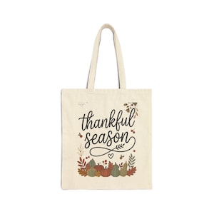 Peut inclure: Un sac fourre-tout en toile beige avec le texte cursif noir "thankful season" centré sur le devant. Le motif comprend une illustration décorative de diverses citrouilles, feuilles et baies aux couleurs automnales le long du bord inférieur.