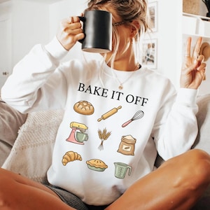 May include: A white long-sleeve sweatshirt featuring the black text BAKE IT OFF and colorful illustrations of baking items including a loaf of bread, rolling pin, whisk, stand mixer, wheat stalks, flour sack, croissant, pie, and measuring cup.