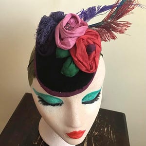 Op de afbeelding: Een zwarte fluwelen fascinator versierd met stoffen bloemen in roze, rode en paarse tinten, geaccentueerd met groene stoffen bladeren en een bos lange, dunne, veelkleurige veren die vanaf de zijkant van het hoofddeksel uitsteken.