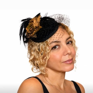 May include: A black fascinator hat featuring a textured base, a decorative brown fabric flower, black feathers, and a delicate black netting veil that partially covers the front. The headpiece is secured to the side of the head.