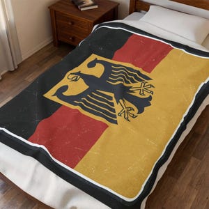 Könnte beinhalten: Eine rechteckige Wohndecke mit einem Used-Look-Druck der deutschen Flagge, bestehend aus den charakteristischen horizontalen Streifen in Schwarz, Rot und Gold sowie einem zentralen schwarzen Adleremblem, umrandet von einem weißen Rand.
